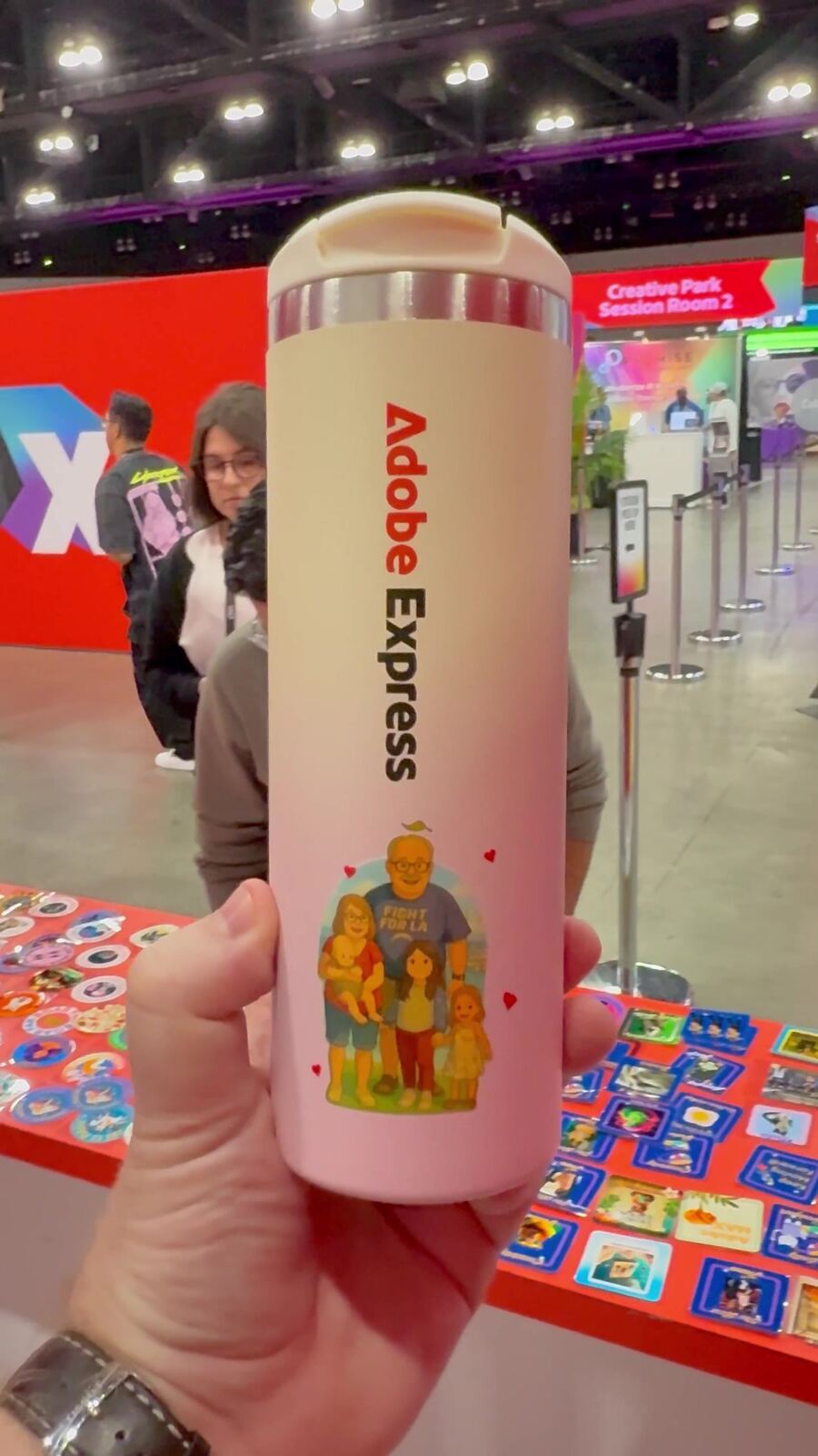 Custom illustrated tumbler held up in front of a busy convention sticker table