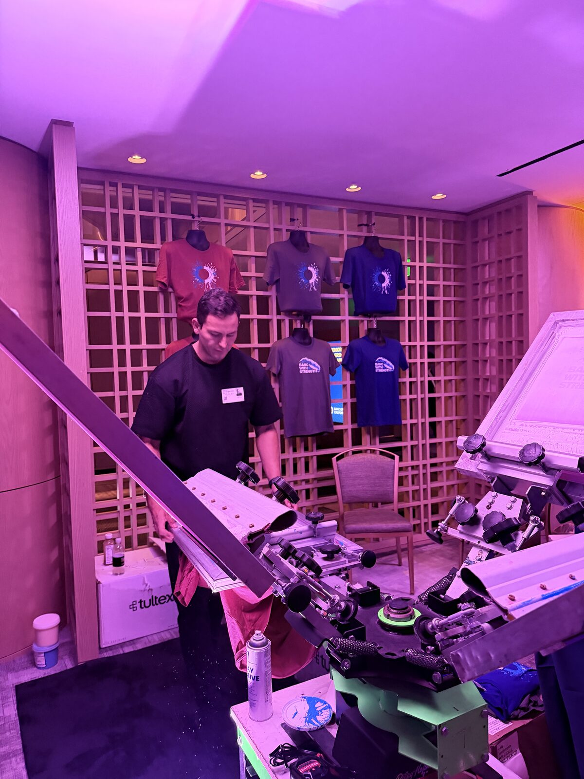 Operator preparing a garment at a live print station inside a hospitality suite with a shirt display wall